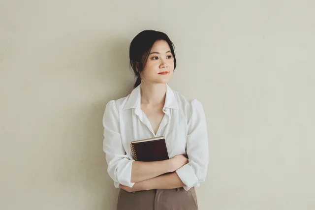 Lauren 穿著白襯衫抱著書，倚牆望向窗光